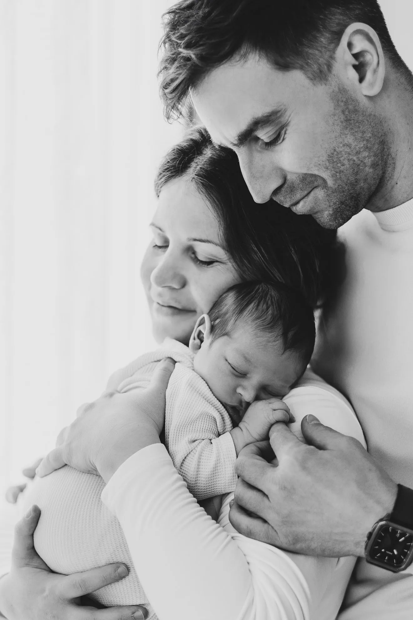 Eine Mutter hält ihr neugeborenes Baby an sich gedrückt an die Schulter und der Vater steht direkt dahinter und berührt das Neugeborene zärtlich am Finger