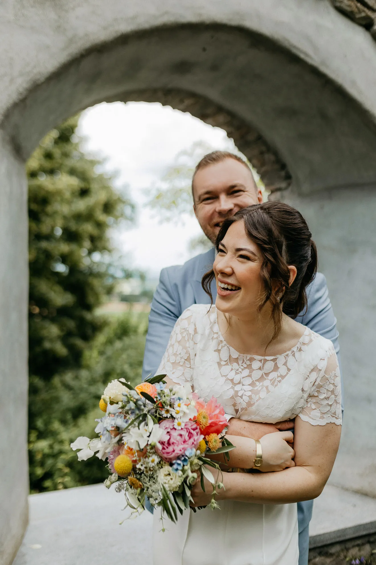Eine Braut läuft vor ihrem frisch angetrauten Ehemann durch einen Torbogen im Bayerischen Wald und hält einen Brautstrauß in der Hand, während er den Arm um ihren Bauch legt