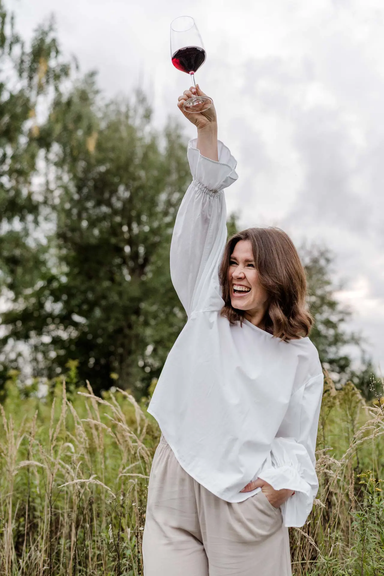 Die Fotografin Katja Stumbeck steht draußen in der Natur vor hohen Gräsern und hält ein mit Rotwein gefülltes Glas in die Luft