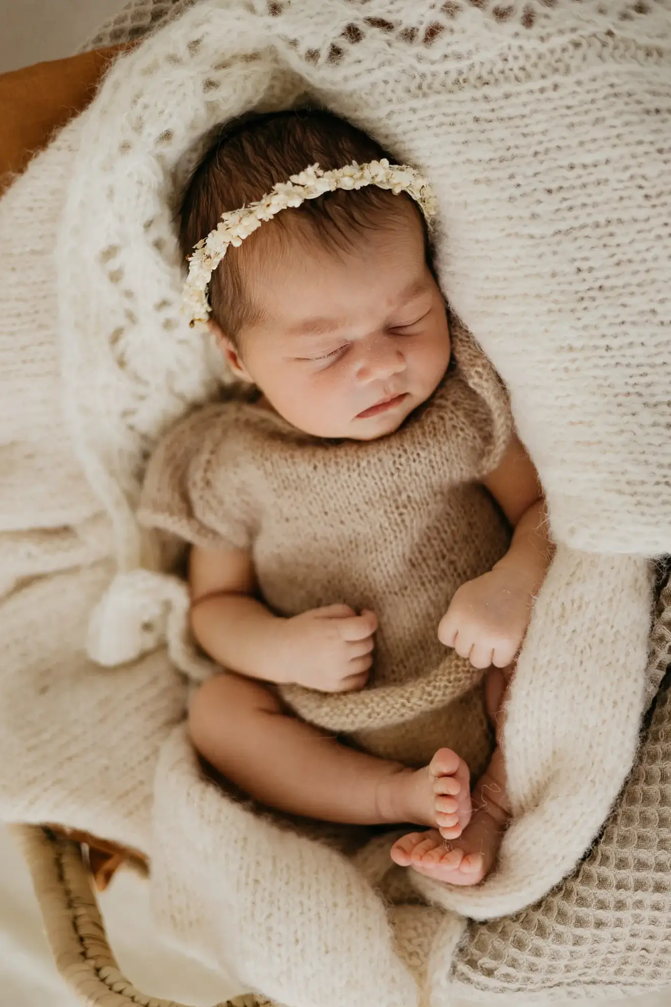 Ein neugeborenes Baby in einem braunen Strickkleid und mit einem Haarkranz aus Blumen liegt zusammengekuschelt in einer weißen Decke und schläft