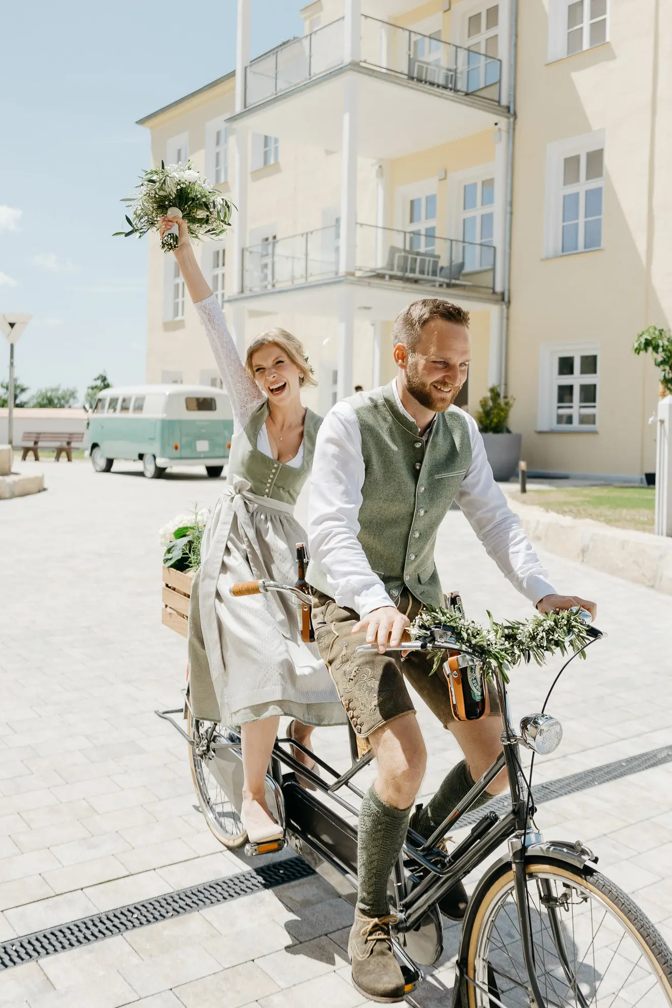 Ein Bräutigam sitzt vorne auf einem Tandemfahrrad und dahinter sitzt seine Braut, die glücklich den rechten Arm mit einem Blumenstrauß in der Hand in die Luft hebt