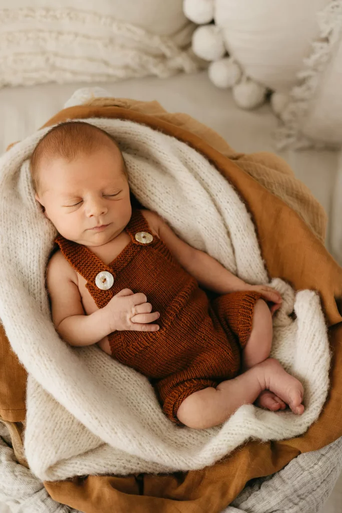 Ein neugeborenes Baby in einer roten Latzhose liegt von mehreren Decken umhüllt auf einer Couch und schläft friedlich