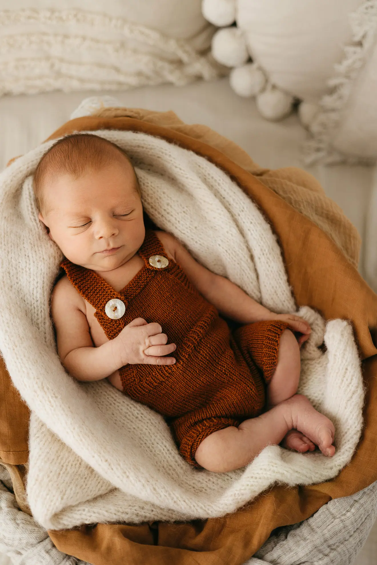 Ein neugeborenes Baby in einer roten Latzhose liegt von mehreren Decken umhüllt auf einer Couch und schläft friedlich