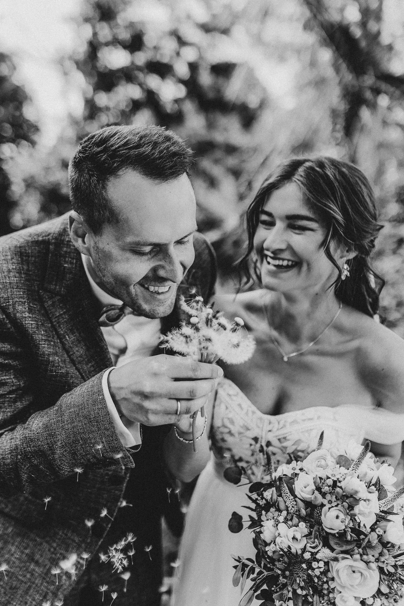 Eine Braut in Offshoulder-Kleid steht neben ihrem Bräutigam und guckt dabei zu wie er einen Strauß aus Pusteblumen zum Fliegen bringt