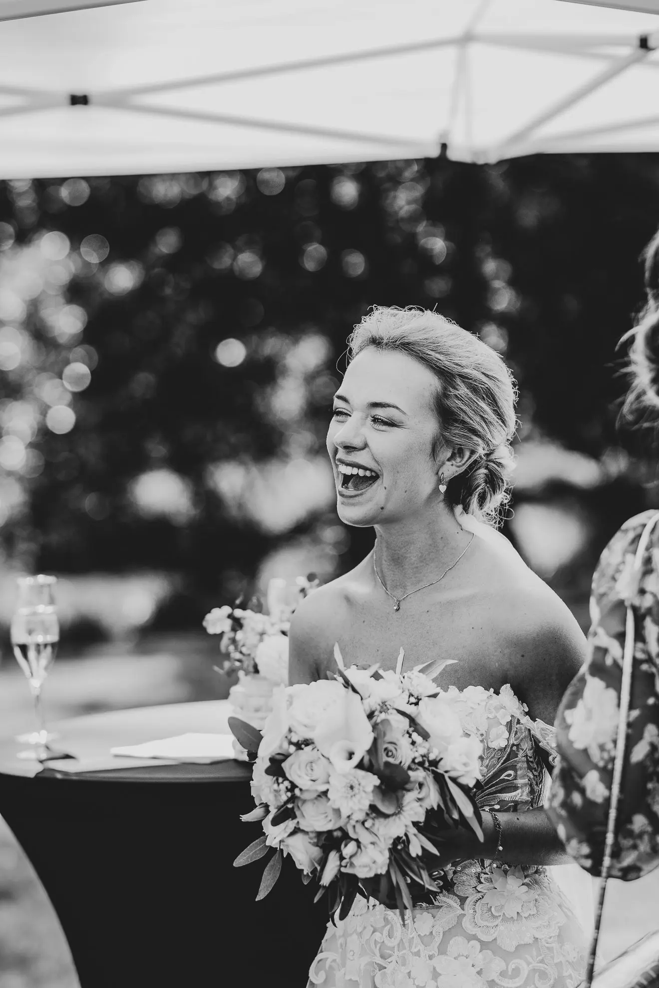 Eine Braut mit zurück gestecktem Haar und Offfshoulder-Kleid strahlt übers ganze Gesicht als die Hochzeitsfotografin Katja Stumbeck eine Schwarz-Weiss-Aufnahme von ihr macht