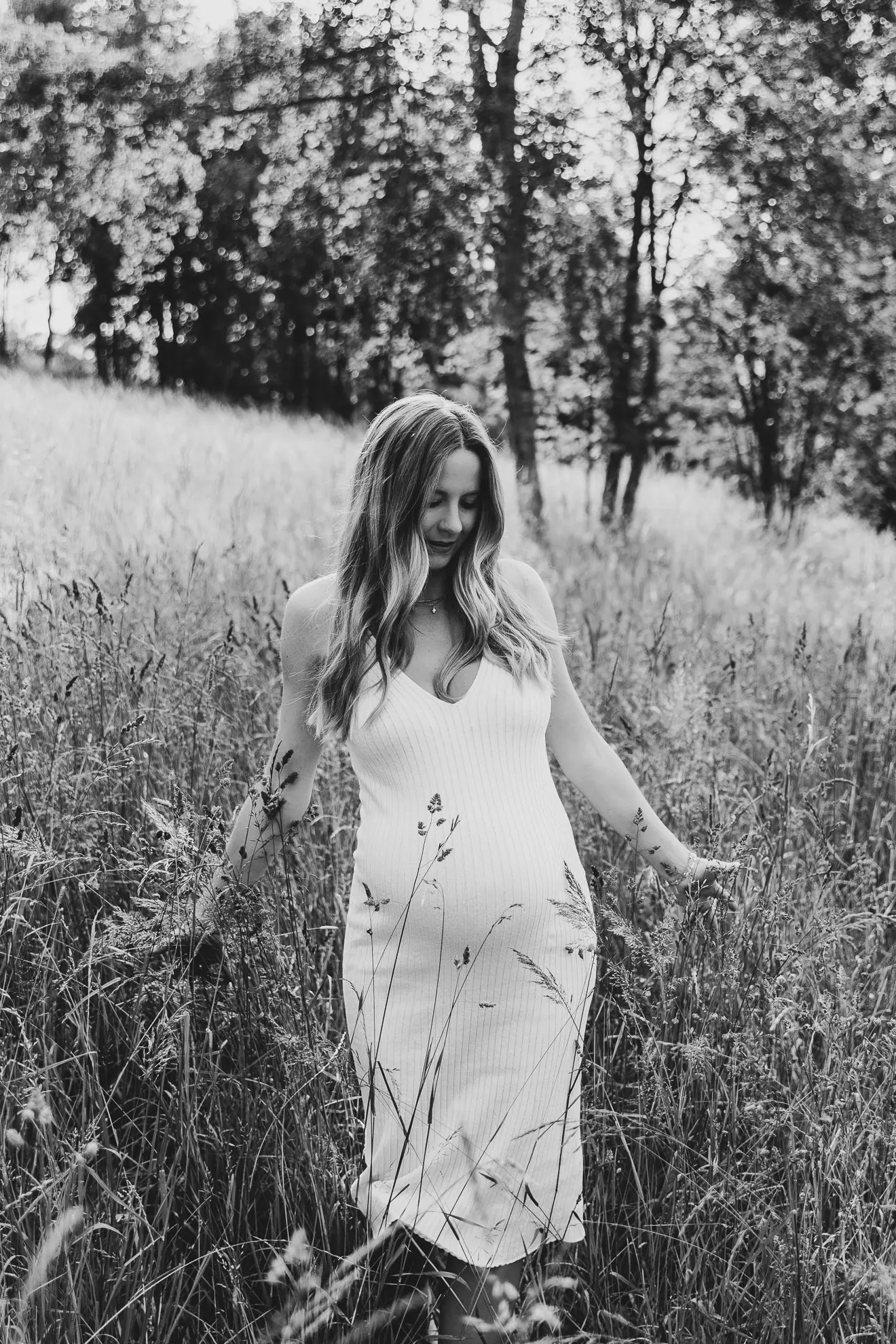 Eine schwangere Frau in einem weißen Kleid mit langen Haaren läuft durch eine Wiese mit hohen Gräsern im Bayerischen Wald und genießt ihre Schwangerschaft