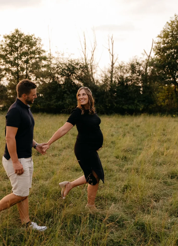 Eine schwangere Frau in einem schwarzen Kleid mit deutlich erkennbarem Babybauch läuft mit ihrem Mann an der Hand über eine Wiese im Bayerischen Wald und strahlt ihn an