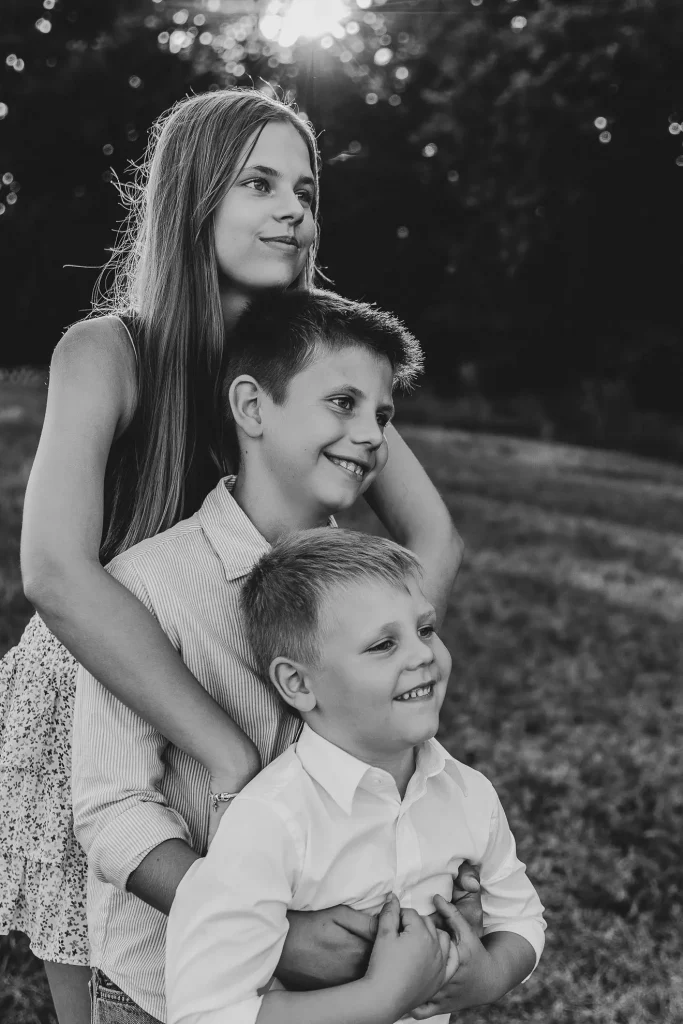 Drei Geschwister, zwei Buben und ein Mädchen, steht übereinander drappiert auf einer Wiese im Bayerischen Wald und werden von Fotografin Katja Stumbeck lächeln in schwarz-weiss fotografiert