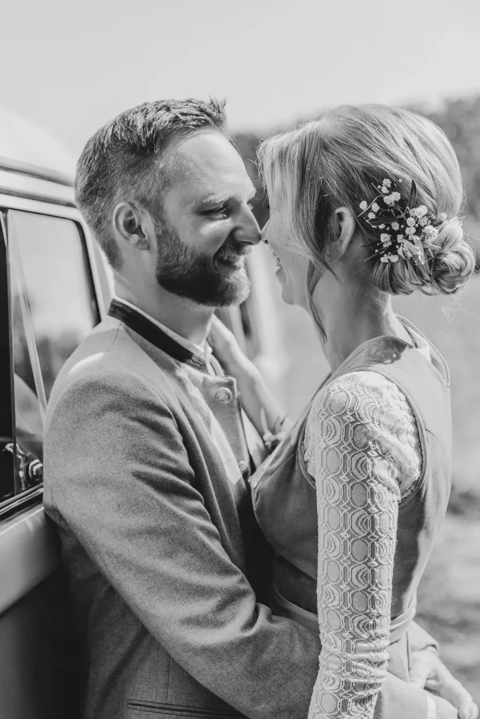 Ein Mann und eine Frau tragen beide Tracht und stehen ganz nah beieinander als sie sich vor ihrem Hochzeitsauto in Straubing tief in die Auge schauen